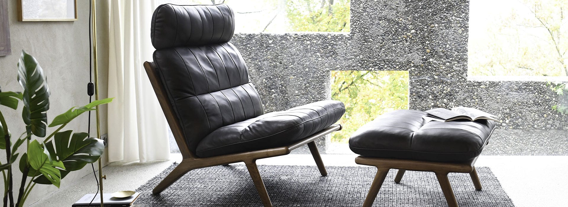 Ein schwarzer Ledersessel mit passender Ottomane steht auf einem dunklen Teppich in der Nähe eines großen Fensters. Auf der Ottomane liegt ein aufgeschlagenes Buch, und neben dem Sessel ist eine Blattpflanze zu sehen. Natürliches Licht fällt von draußen herein.