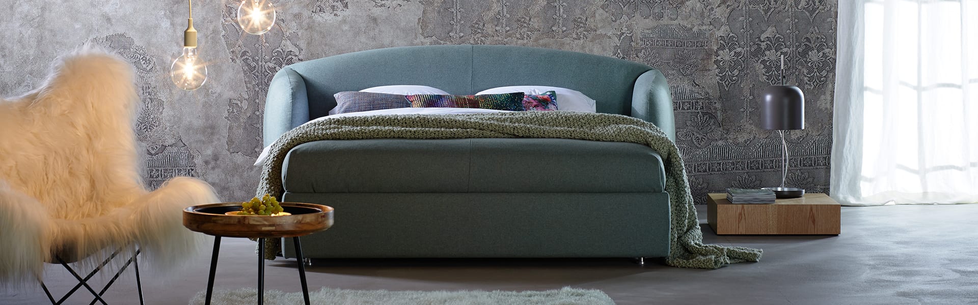 Ein modernes Schlafzimmer mit einem grün gepolsterten Bett, einer strukturierten grauen Wand, einem Beistelltisch mit einer Lampe, einem kleinen runden Tisch mit Dekoration und einem weißen Plüschsessel. Pendelleuchten hängen von der Decke und das Sonnenlicht fällt durch einen durchsichtigen Vorhang.