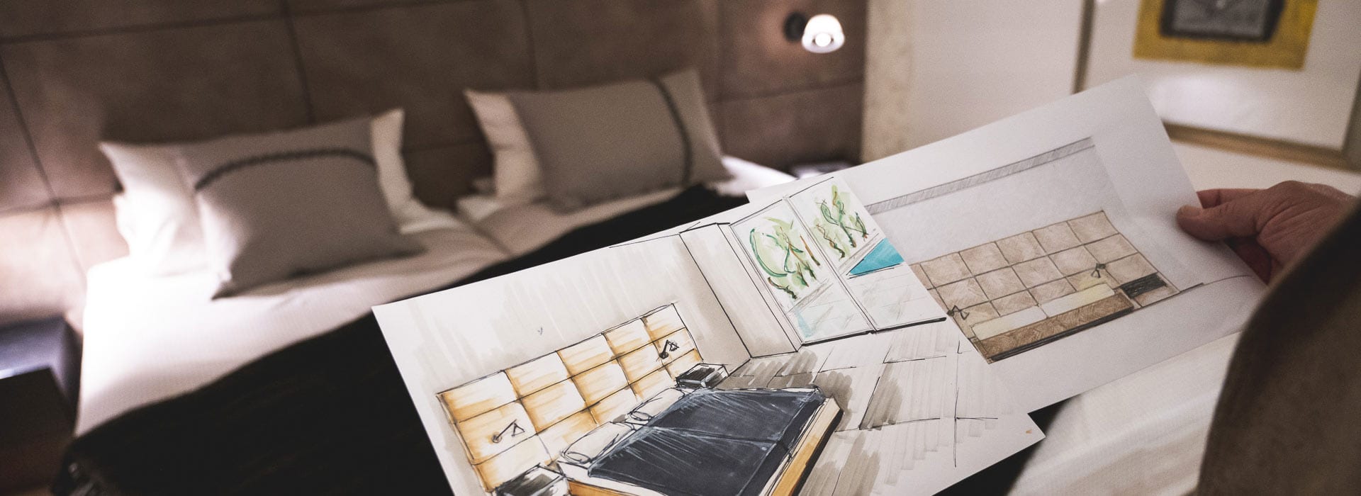 Eine Person hält Einrichtungsskizzen in der Hand, die genau zu dem modernen Schlafzimmer im Hintergrund passen, das mit einem Doppelbett mit neutraler Bettwäsche, Wandlampen und einem braunen gepolsterten Kopfteil ausgestattet ist.