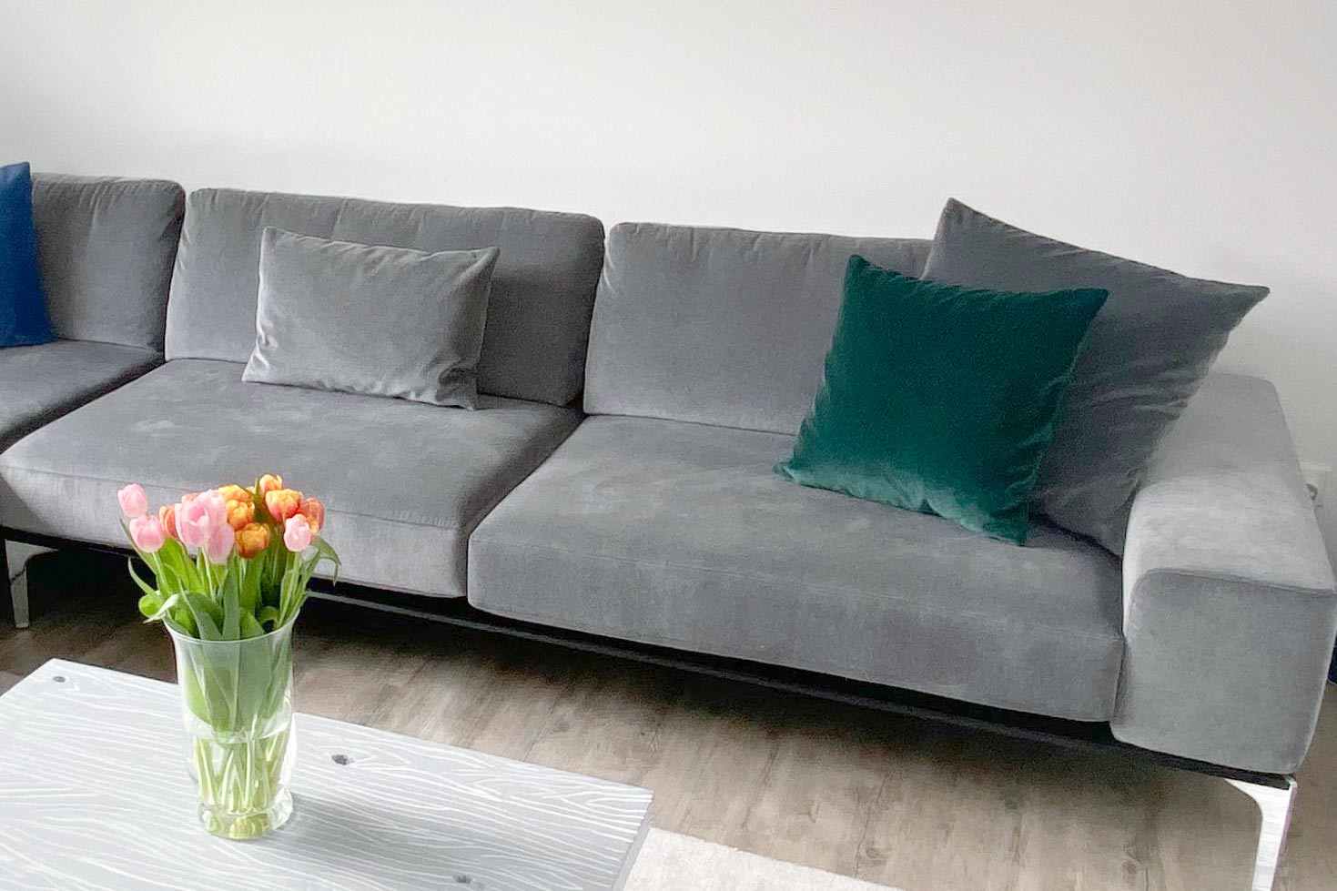Ein graues Sektionssofa mit zwei Kissen, einem grauen und einem dunkelgrünen, steht vor einer weißen Wand. Vor dem Sofa steht ein heller Tisch mit einer Vase mit rosa und orangefarbenen Tulpen. Der Boden ist aus Holz.