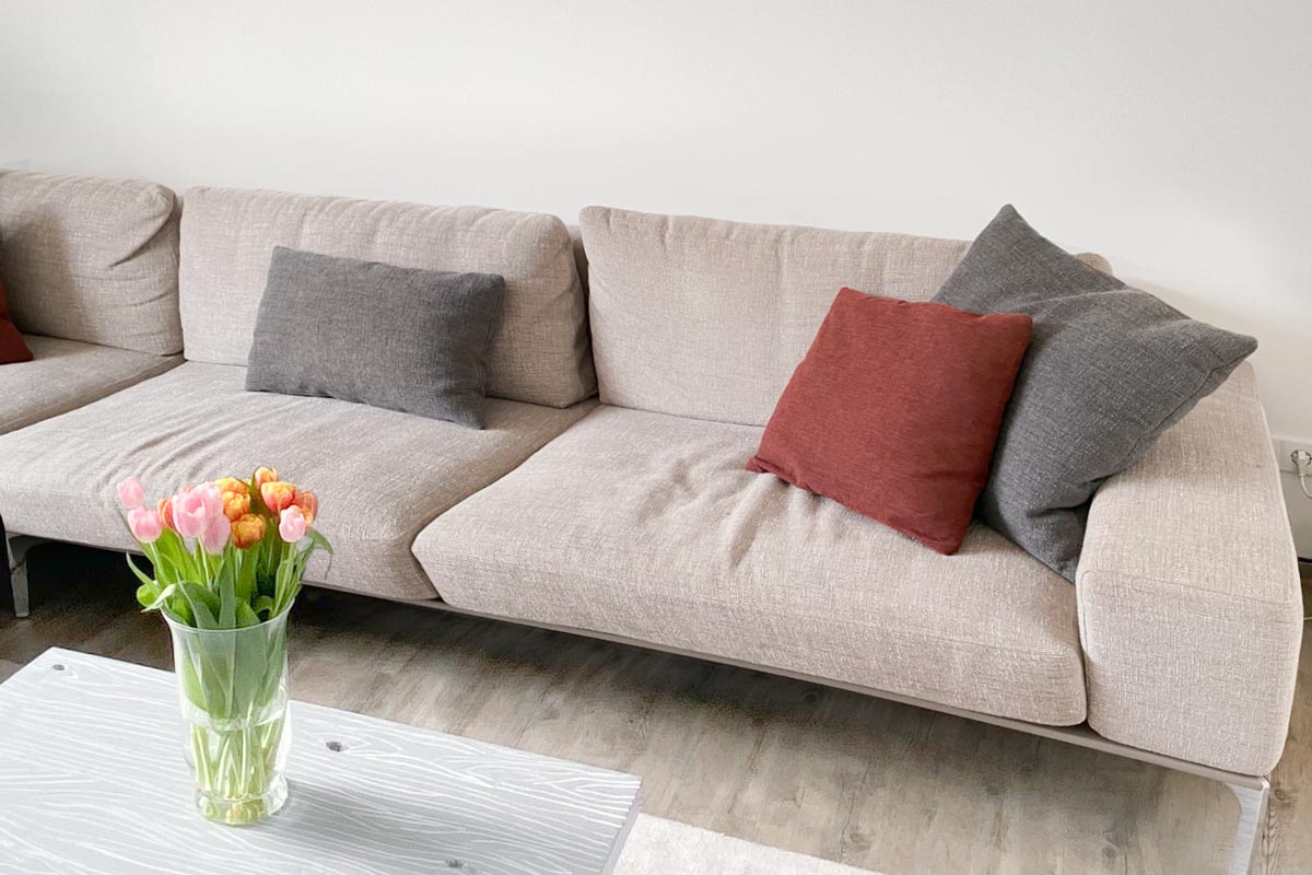 Ein beiges Sektionssofa mit zwei grauen und zwei roten Wurfkissen steht an einer weißen Wand. Davor steht eine Glasvase mit rosa und orangefarbenen Tulpen auf einem hellen Couchtisch.